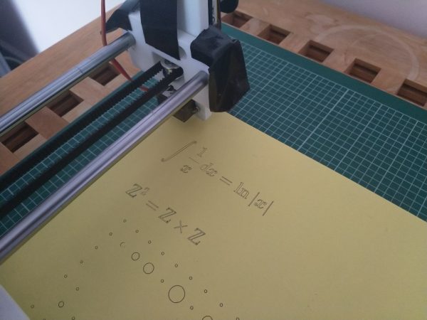 The pen plotter hovering over a piece of paper, on which is drawn some mathematical notation. Each of the symbols has been drawn in outline.
