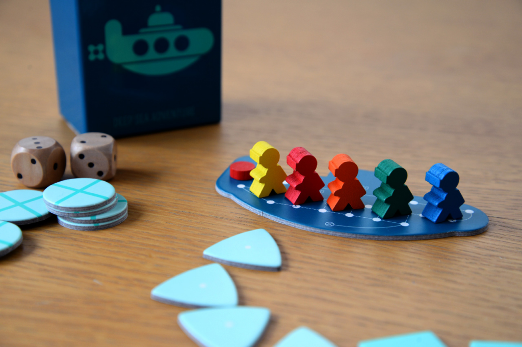 A photo of board game pieces on a wooden table in front of the game box, including round tokens, triangular tokens, person-shaped wooden pieces and two dice