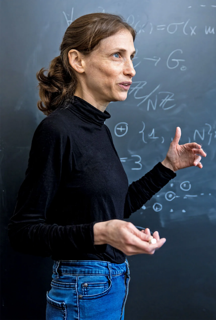A photo of Rachel Greenfield, a white female mathematician, wearing a polo neck and jeans and talking in front of a blackboard