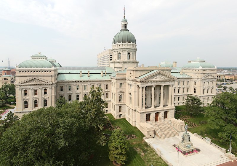 Photo of the Indiana Statehouse, a large building with columns and arched windows, cream walls and green domed roofs, set in a garden in the middle of a city