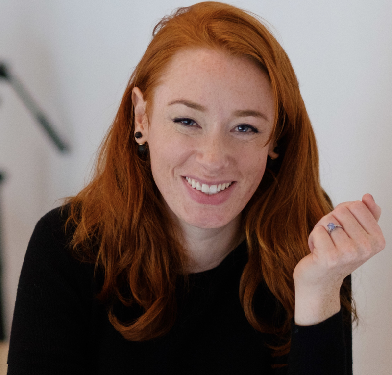A photo of Hannah Fry, a young woman with long red hair, smiling while wearing a black top, with one elbow resting on a table