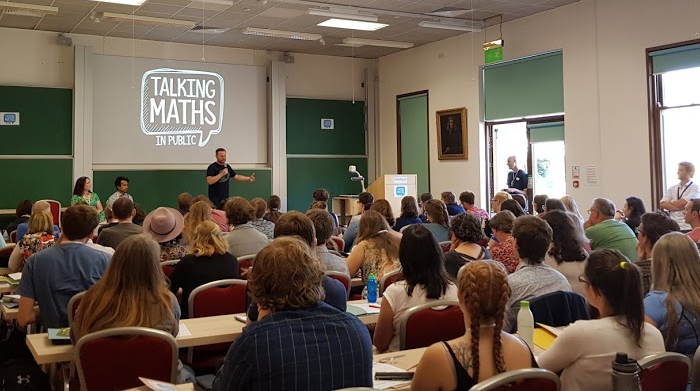 Photo of Brady, Anand and Jen in front of the audience in a lecture theatre, with the "Talking Maths in Public" logo on the screen behind them