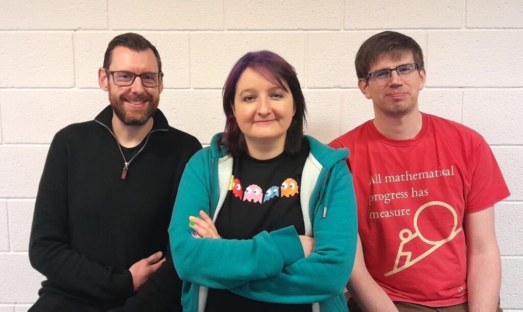 Photo of Peter, Katie and Christian standing in front of a white brick wall. Peter is a white man with glasses, a beard and short brown hair; Katie is a white woman with purple hair and a pac-man t-shirt; Christian is a white man with short brown hair and glasses and a maths t-shirt on