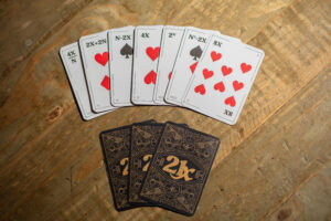A spread of 21X cards on the table, including 4X/N, 2X+2N, N-2X, 4X, 2^N etc. Some face-down cards are below, and the backs have a dark design with an intricate gold pattern around the 21X logo.