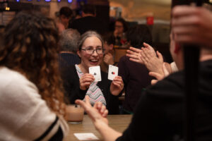 A group of people playing 21X. An older white woman with glasses holds up two cards, while others applaud.