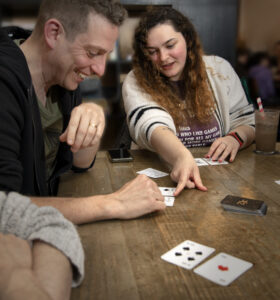 People sitting around a table playing 21X. A long-haired white woman points to a card on the table, while a smiling white man looks on.