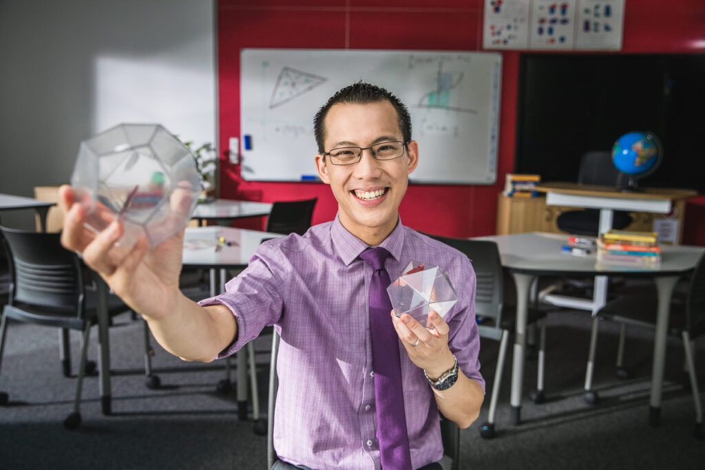 Promotional photo for Eddie's channel. He's wearing a purple shirt, standing in a math classroom and holding up some clear plastic polyhedra.