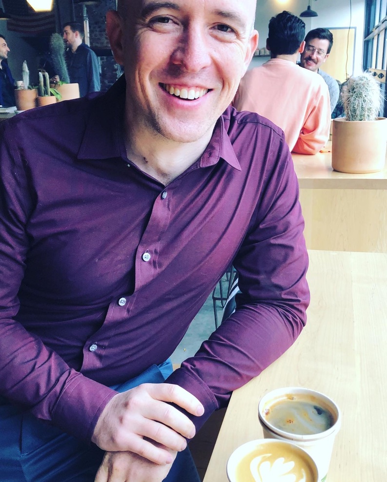 A photo of Keenan Crane, a smiling white man in a purple shirt leaning on a bar next to a cup of coffee