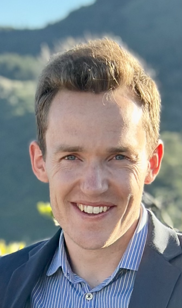 Photo of Grant Sanderson in front of a hill; he is a white man with short brown hair and a tall narrow face, who is wearing a shirt and smiling