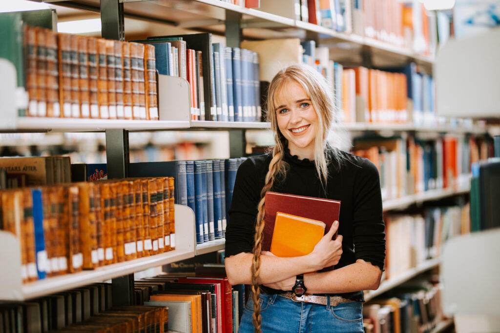 A photo of Toby, a young white woman with long blonde hair in a plait wearing a black top and jeans, standing in a library next to a shelf of maths books holding two books