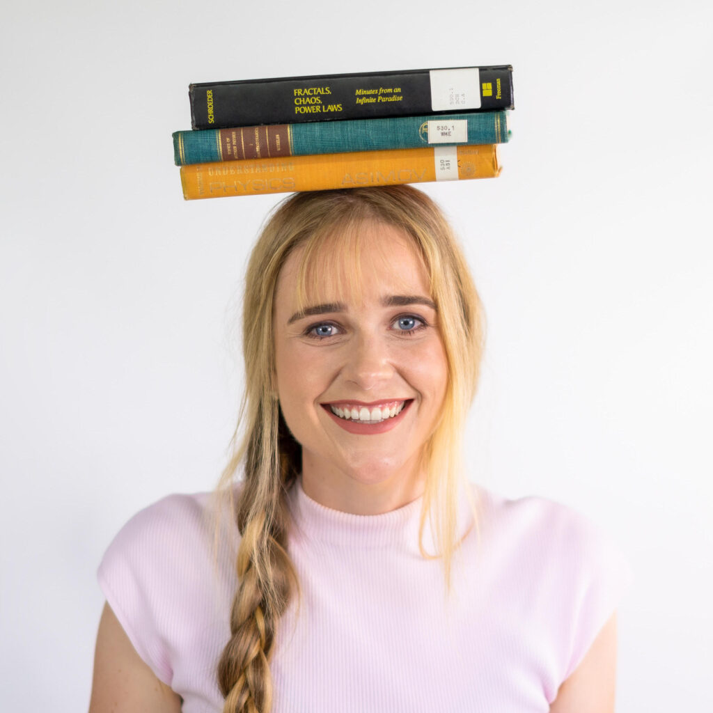A photo of Toby, a young white woman with long blonde hair in a plait wearing a pale pink tshirt, smiling with a pile of maths textbooks balanced on her head
