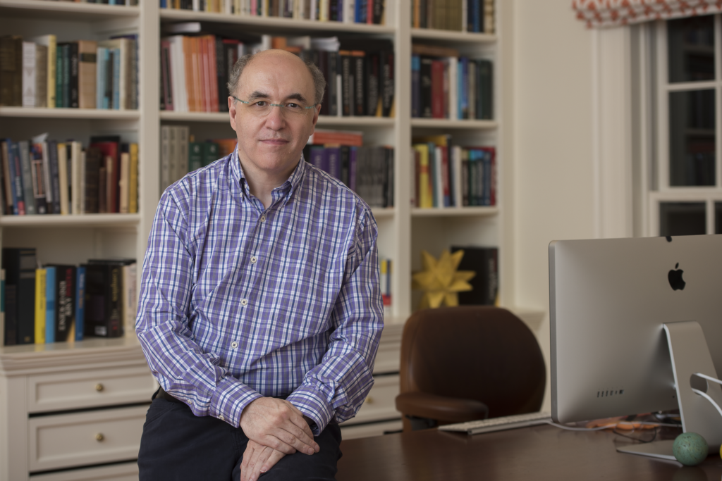 Photo of Stephen Wolfram, a balding white man with glasses and a checked shirt, leaning on a desk in an office