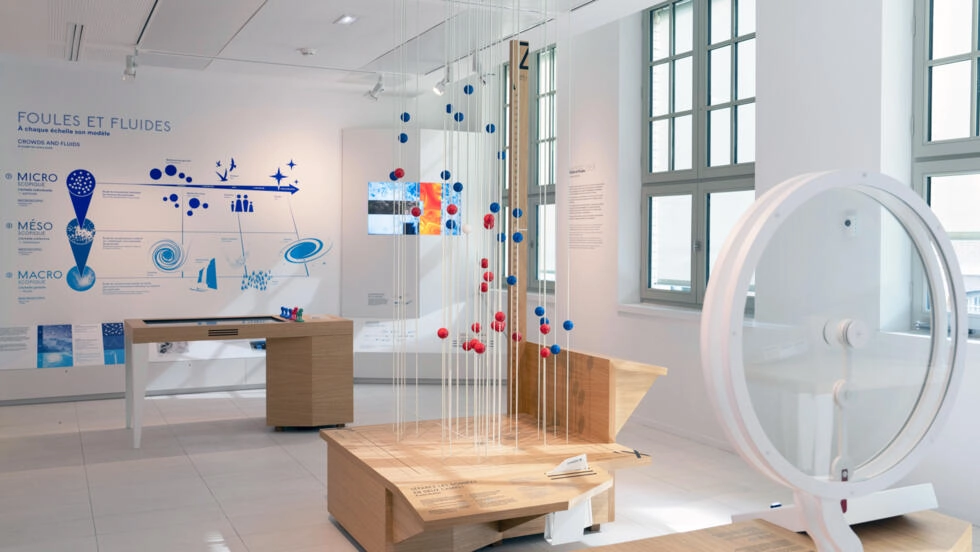 A photo showing some exhibits in the maths museum, including a double pendulum, a set of balls arranged in 3D space on some vertical strings as a plot, and a wall display about fluid dynamics.