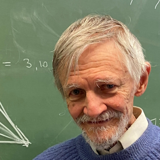 A photo of Miles Reid, a white man with grey hair and a beard, who is looking coy in front of a blackboard