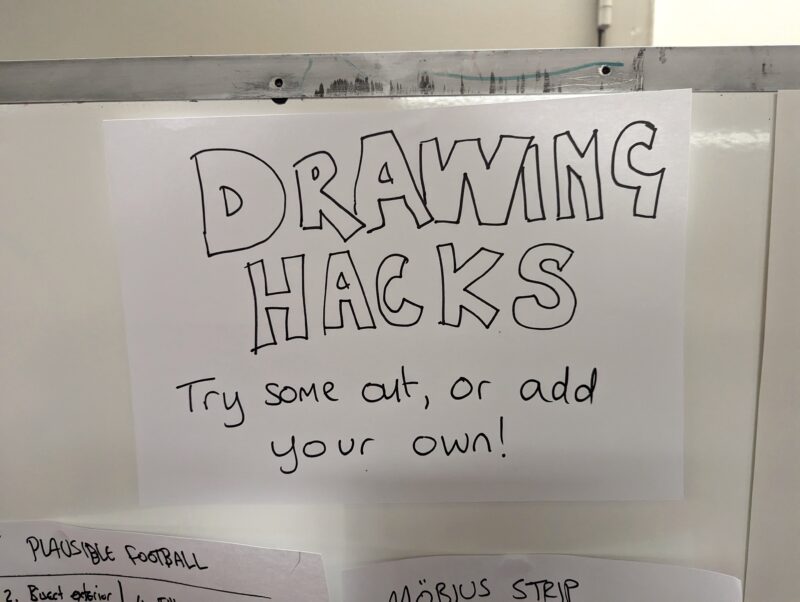 Photo of the header of our board of 'Drawing Hacks' - a piece of paper attached to a whiteboard which says 'Drawing Hacks - Try some out, or add your own!'