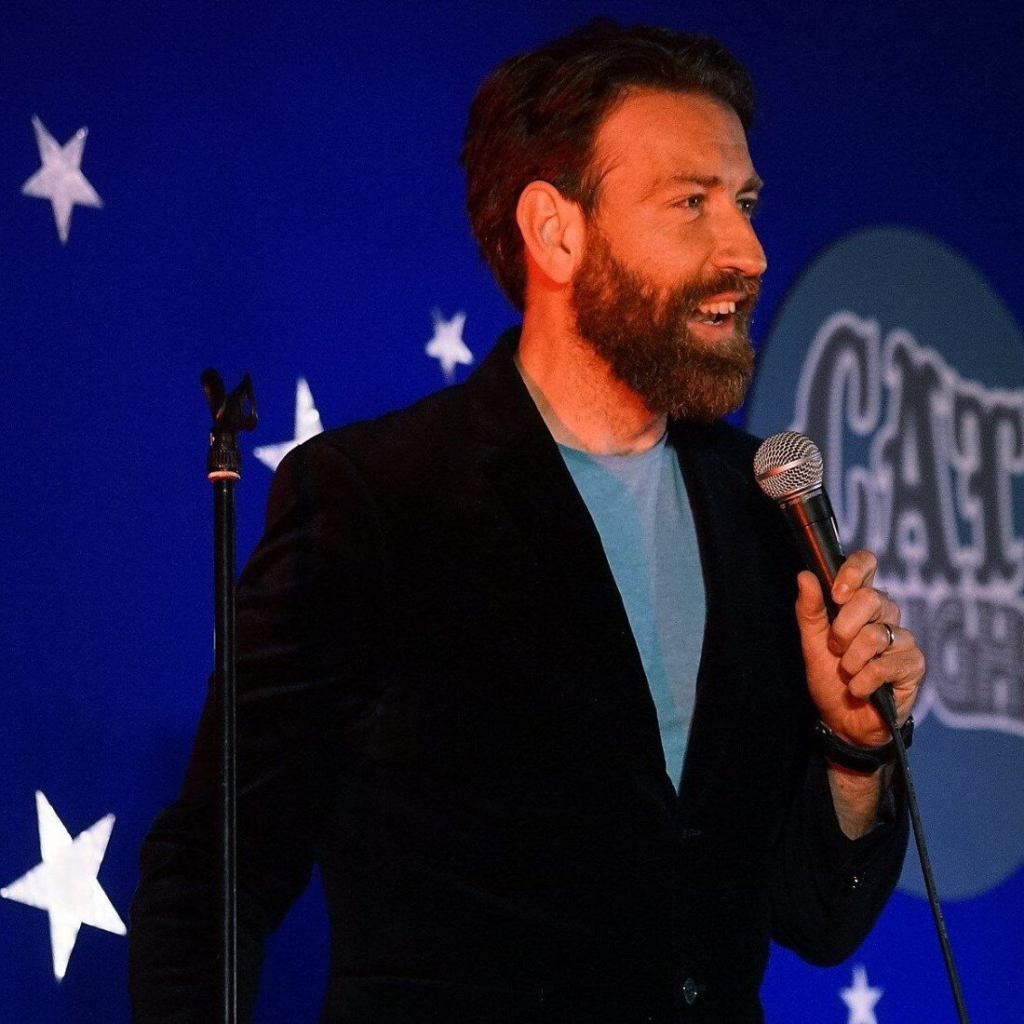 Photo of Colm O'Regan, a white man with short brown hair and a beard, talking into a microphone on stage at a comedy show