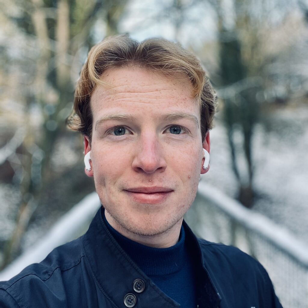 Photo of Sam, a young white man with short ginger hair, looking into the camera while wearing Apple earbuds and a dark blue top
