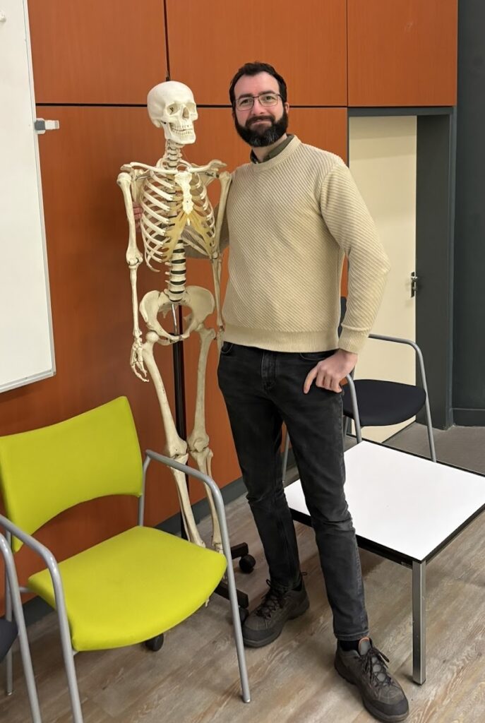 Photo of Marcello Seri, a tall white man with black hair and a full beard, wearing a beige jumper and black jeans, smiling and standing next to a skeleton
