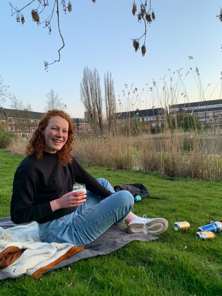 Photo of Marit, a young woman with long curly red hair, sitting having a picnic, smiling and wearing jeans and a black jumper