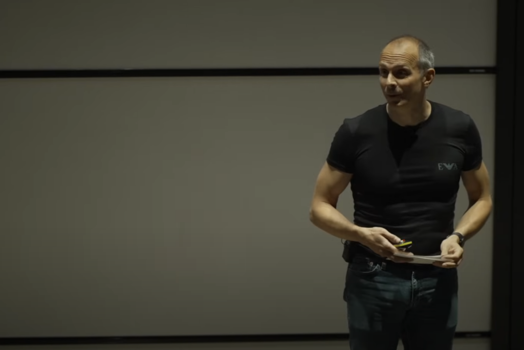 Screenshot of the video of Tim Harford giving the inaugural Vicky Neale Memorial Lecture; a balding white mand in a t-shirt and jeans is standing on the right in front of a projector screen