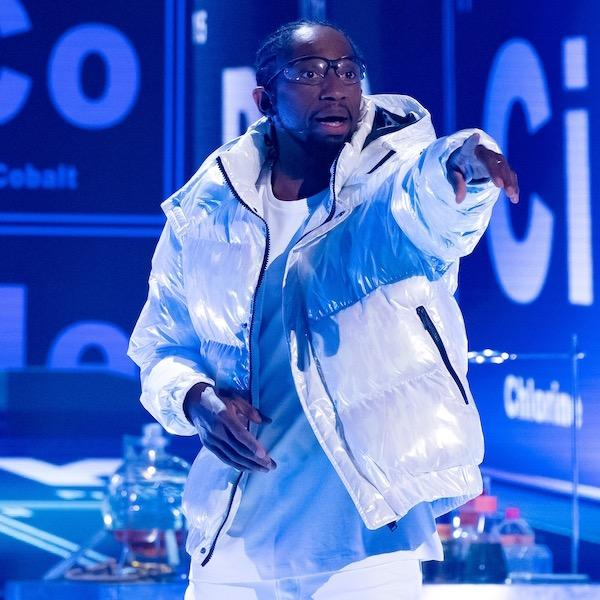 Photo of Jon, a black man with his hair in cornrows, wearing a white puffer jacket and rapping in front of a stage decorated with chemical element symbols