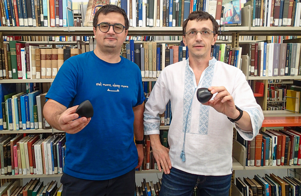 Photo of two white men with short brown hair and glasses, each holding a black solid of constant width (a rounded tetrahedron). One is wearing a traditional Ukrainian embroidered shirt