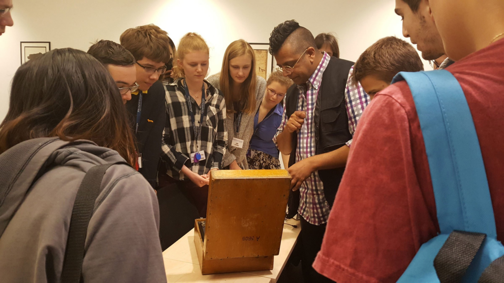 Photo of a collection of teenage students (roughly half boys and half girls), crowded around looking at an Enigma machine, which looks like an open wooden box on a table, with Simon Singh, an asian man with a mohican and glasses wearing a gilet over a check shirt