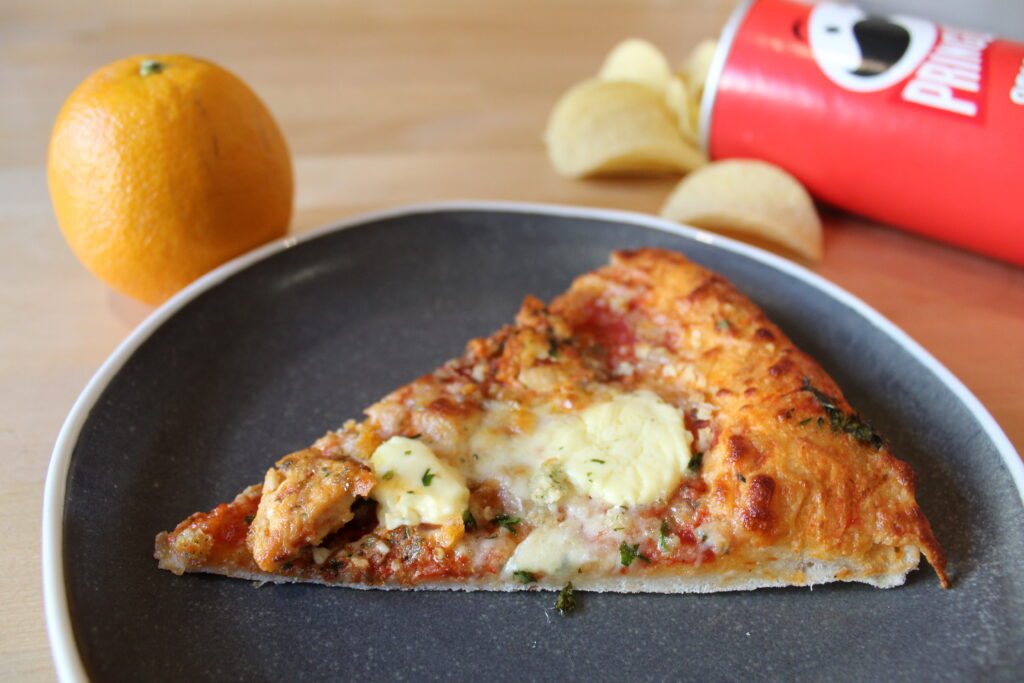Slice of pizza on a plate next to an orange and some Pringles.