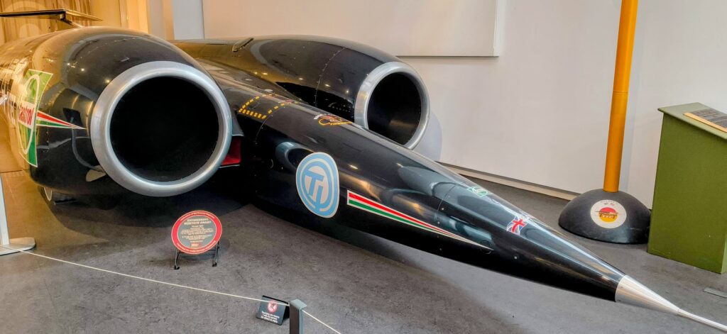 The sleek, black Thrust SSC land-speed record-holder stretches across a museum gallery, its needle-like nose tapering to a sharp silver point while two enormous jet engine intakes loom behind the cockpit like giant circular tunnels. The machine’s glossy body is decorated with racing logos, thin red-white-green stripes, and a small Union Flag near the front, emphasising its British engineering heritage. Low barriers and information plaques surround the exhibit, while the dramatic proportions - extremely long, low, and narrow - make the car look more like an aircraft fuselage or missile than a conventional car, hinting at the extraordinary speeds it was built to achieve.