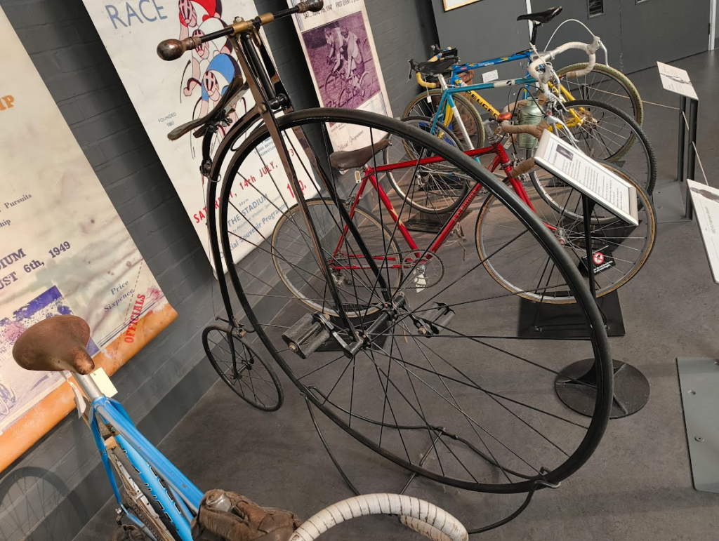 A black metal-framed penny-farthing bicycle dominates a museum display of historic cycles, its enormous front wheel rising above the surrounding exhibits while the tiny rear wheel almost disappears behind it. The polished metal frame and high saddle emphasise just how precarious riding it must have felt. Around it, several later racing bicycles in bright reds and blues are arranged on stands, with vintage cycling posters and information panels lining the walls, creating a timeline of cycling history from the Victorian era onwards.
