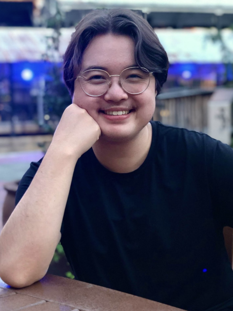 Photo of Stan, a young asian man with round glasses and mid-length dark hair. Stan is smiling and resting his chin on his closed fist, with an elbow on the table.